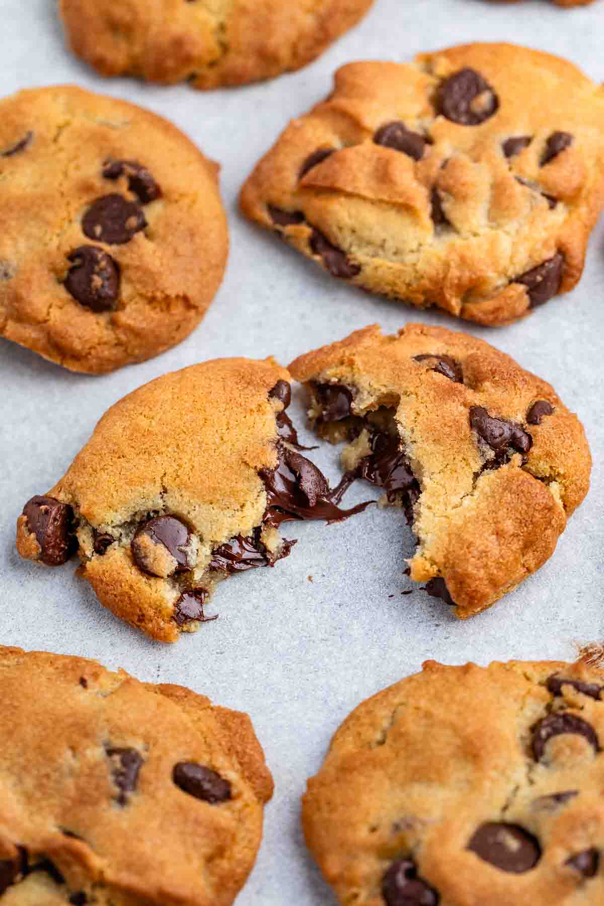 Close-up of chocolate chip cookies on a baking sheet. One cookie is broken in half, showing melted chocolate chips inside. The cookies have a golden-brown crust with visible chocolate chunks.