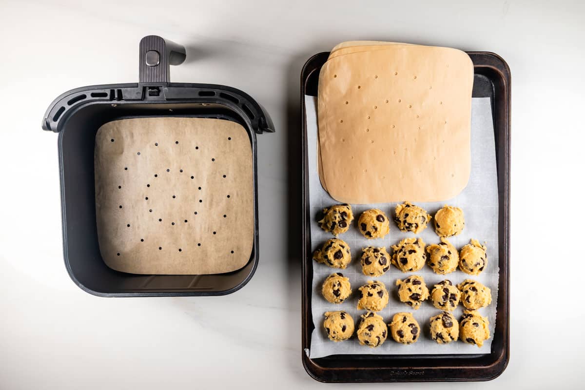 An air fryer basket with a round perforated parchment paper on the left, and a baking tray on the right with a square perforated parchment paper and 18 raw chocolate chip cookie dough balls arranged in rows.