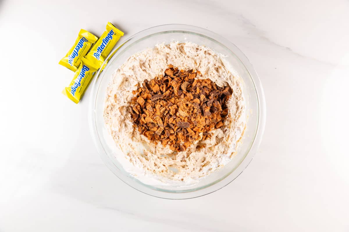 A glass bowl filled with a creamy mixture topped with crushed candy pieces, positioned on a white surface. Three small, yellow Butterfinger candy packages are placed next to the bowl.