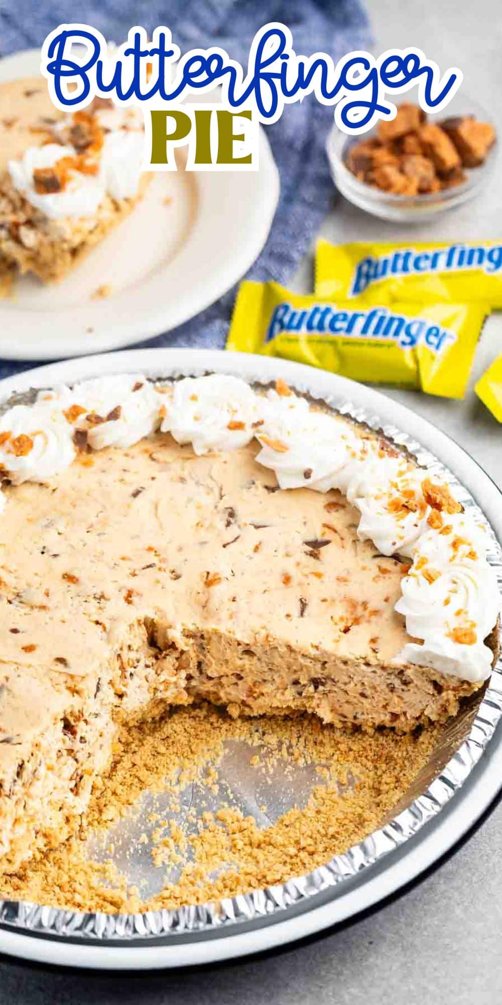 A pie with a graham cracker crust, creamy filling, and pieces of Butterfinger candy is topped with whipped cream. One slice is missing. In the background, there are Butterfinger candy wrappers and a small plate with a slice of pie.