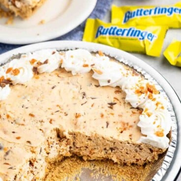 A creamy pie with a graham cracker crust, topped with whipped cream and crushed candy pieces, has a slice removed. A slice is served on a white plate in the background. Three yellow candy wrappers labeled Butterfinger are nearby.