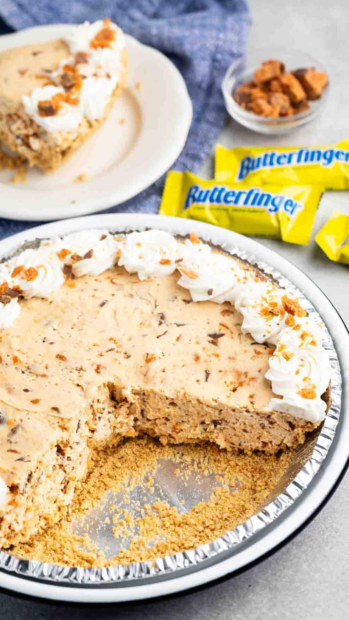 A creamy pie with a graham cracker crust, topped with whipped cream and crushed candy pieces, has a slice removed. A slice is served on a white plate in the background. Three yellow candy wrappers labeled Butterfinger are nearby.