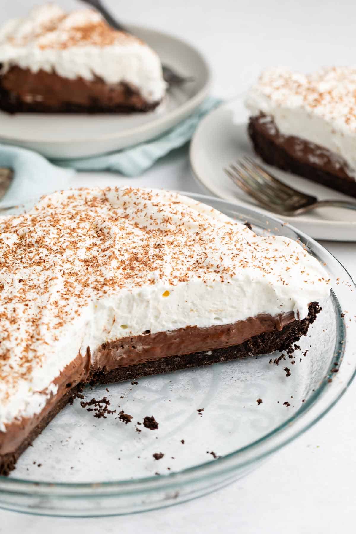 A chocolate cream pie with a slice removed, revealing layers of creamy chocolate filling topped with whipped cream and chocolate shavings. Two plates in the background hold slices of the pie with forks beside them.
