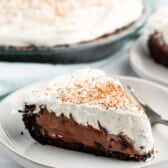 A slice of chocolate cream pie on a white plate with a fork. The pie has a chocolate filling, whipped cream topping, and a sprinkle of cocoa powder. The remaining pie is visible in the background on a glass plate.