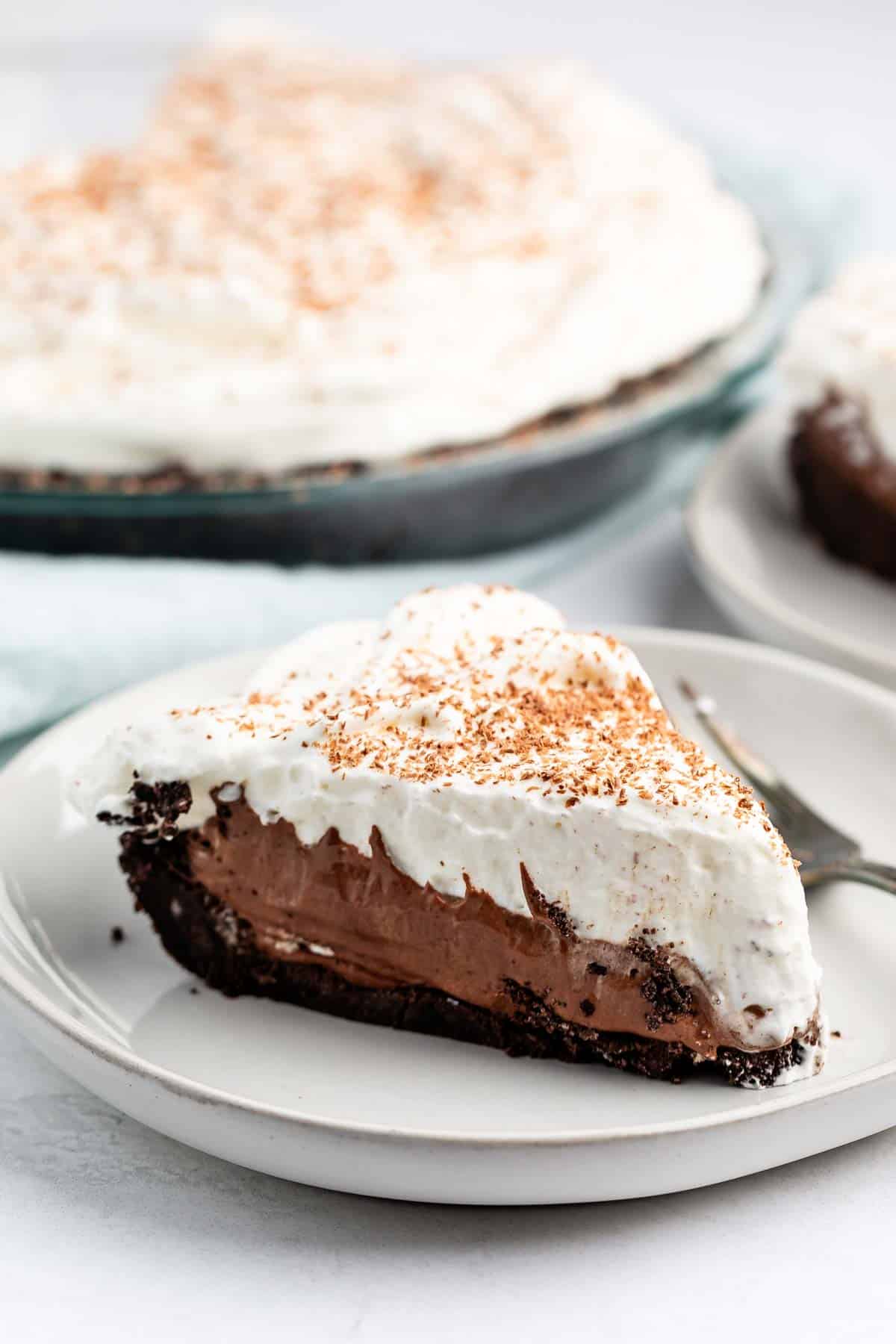 A slice of chocolate cream pie on a white plate with a fork. The pie has a chocolate filling, whipped cream topping, and a sprinkle of cocoa powder. The remaining pie is visible in the background on a glass plate.