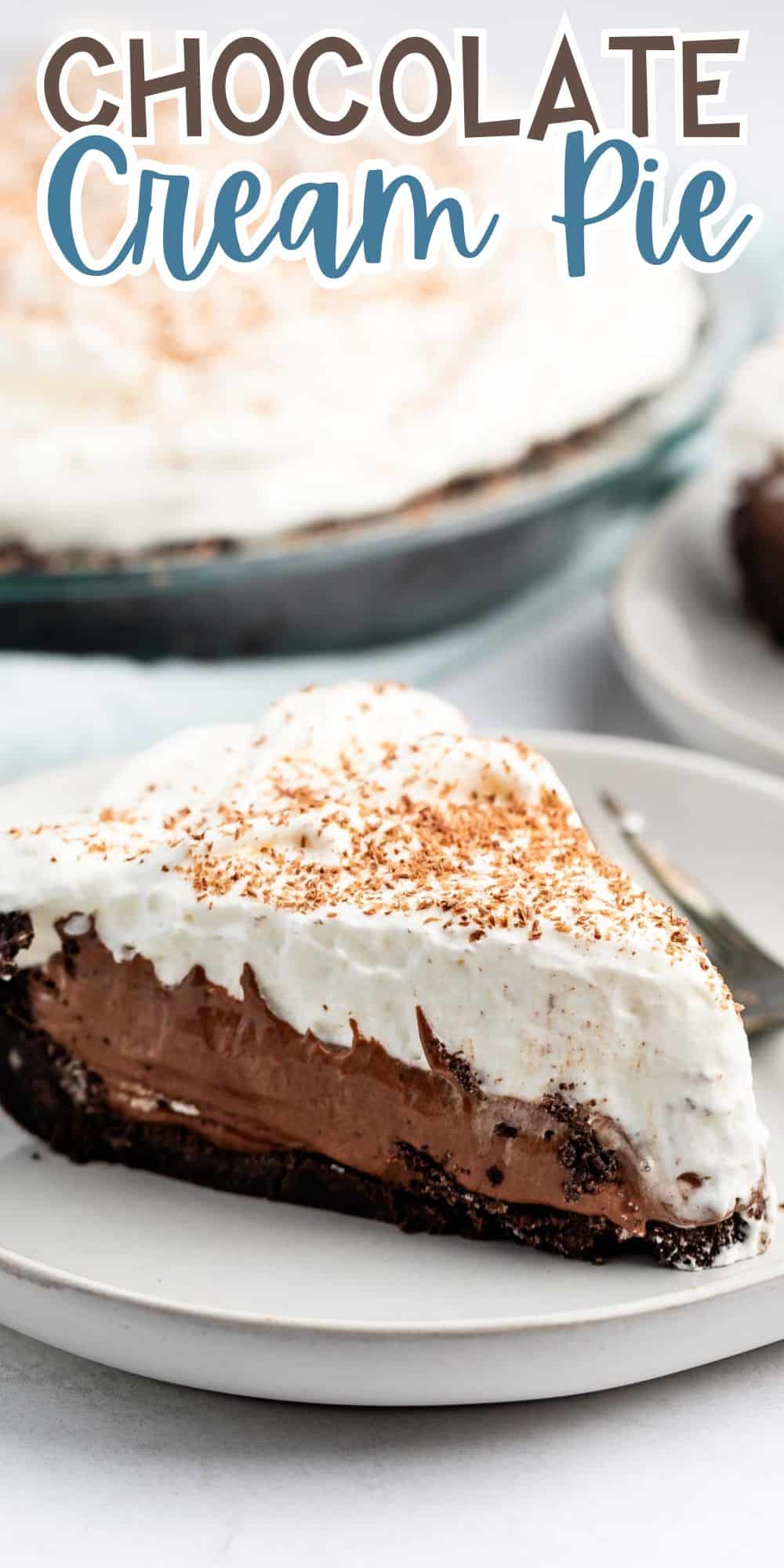 A slice of chocolate cream pie on a white plate with a chocolate crust and whipped cream topping. The pie is garnished with cocoa powder, with more pie visible in the background. A fork is placed beside the slice.