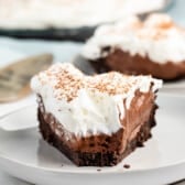 A slice of chocolate pie on a plate, featuring a crumbly chocolate crust, rich chocolate filling, and a thick layer of whipped cream topped with chocolate shavings. A larger pie and a metal server are visible in the background.