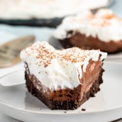 A slice of chocolate pie on a plate, featuring a crumbly chocolate crust, rich chocolate filling, and a thick layer of whipped cream topped with chocolate shavings. A larger pie and a metal server are visible in the background.