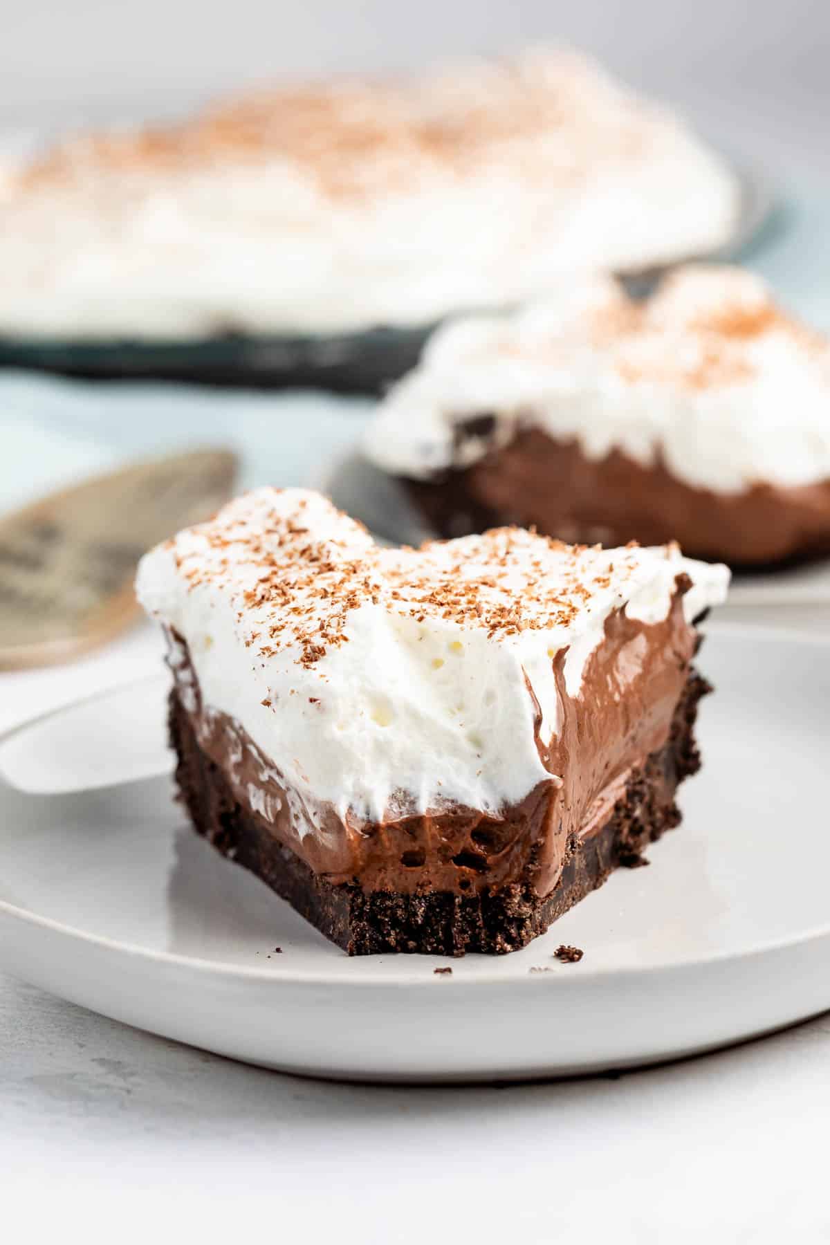 A slice of chocolate pie on a plate, featuring a crumbly chocolate crust, rich chocolate filling, and a thick layer of whipped cream topped with chocolate shavings. A larger pie and a metal server are visible in the background.