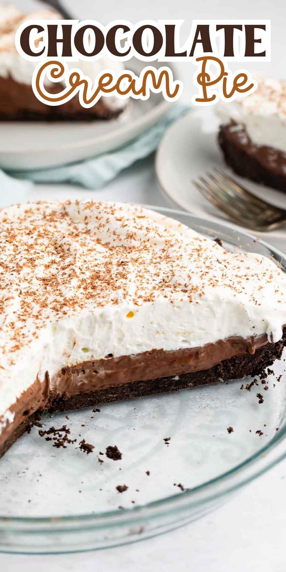 A chocolate cream pie in a glass dish, topped with whipped cream and chocolate shavings. A slice has been removed, revealing a rich chocolate filling and a dark crust. Plates with a slice of pie are blurred in the background.