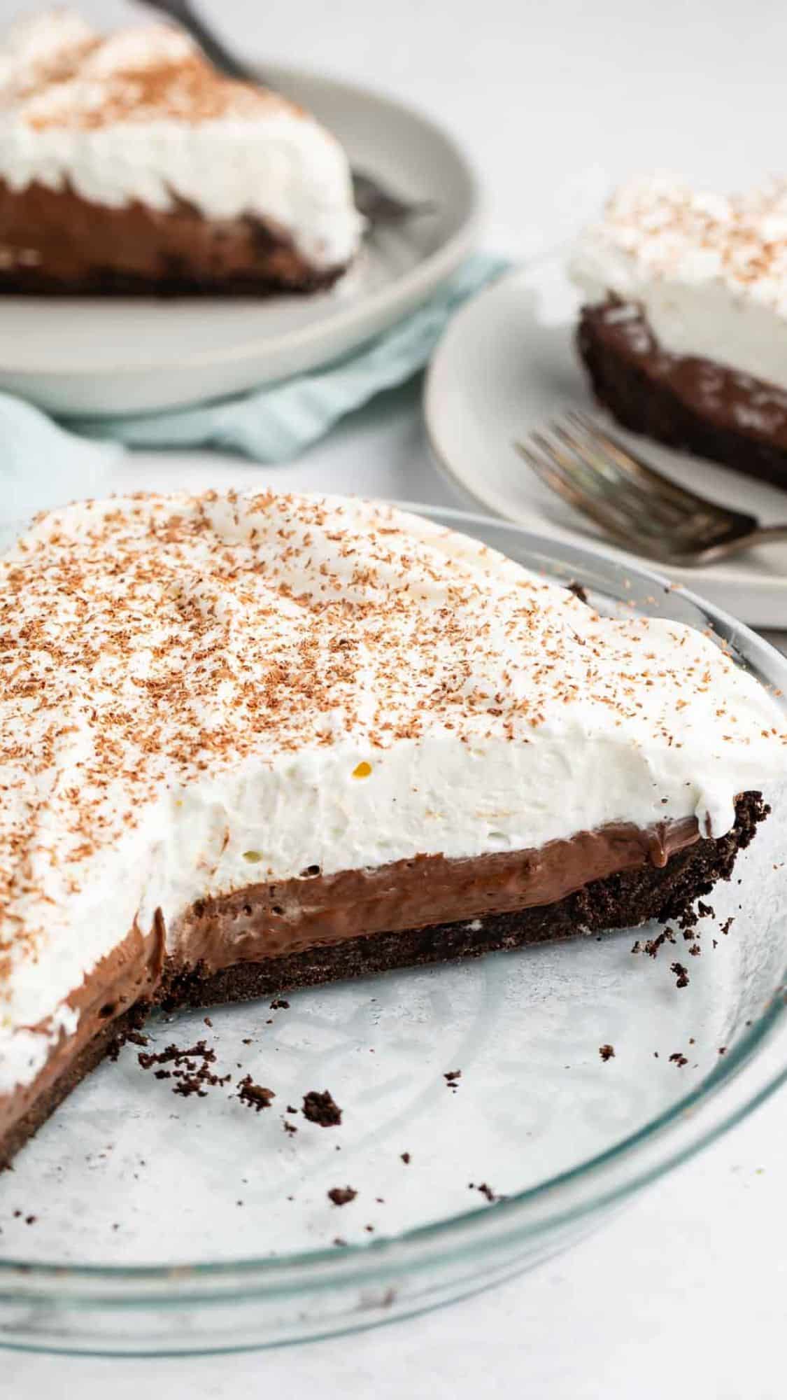 A close-up of a chocolate cream pie in a glass dish. The pie has a rich chocolate filling topped with whipped cream and chocolate shavings. Several slices are already served on plates in the background, each with forks beside them.