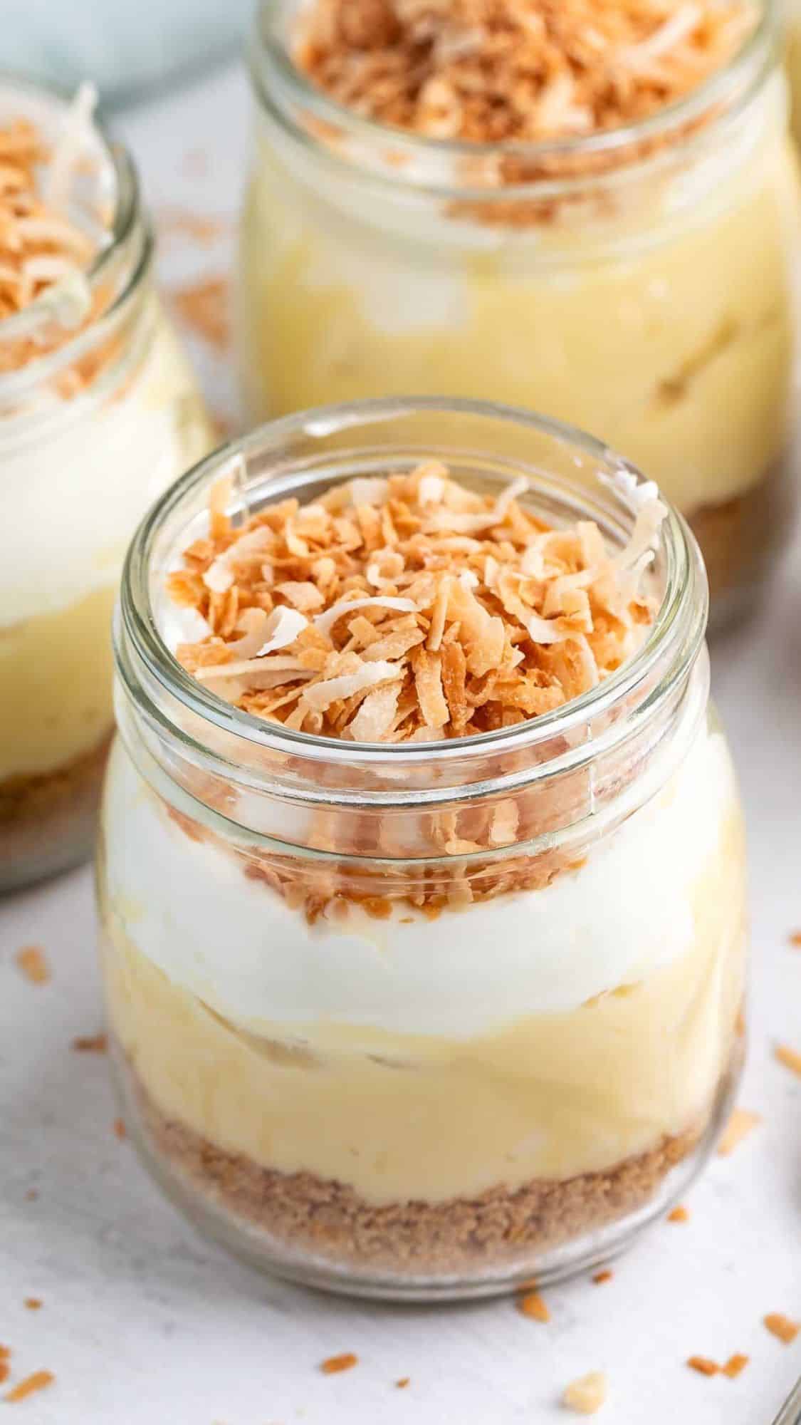 Jar dessert with layers of crumbled crust, creamy filling, and topped with whipped cream and toasted coconut flakes. Multiple jars are in the background on a light surface.