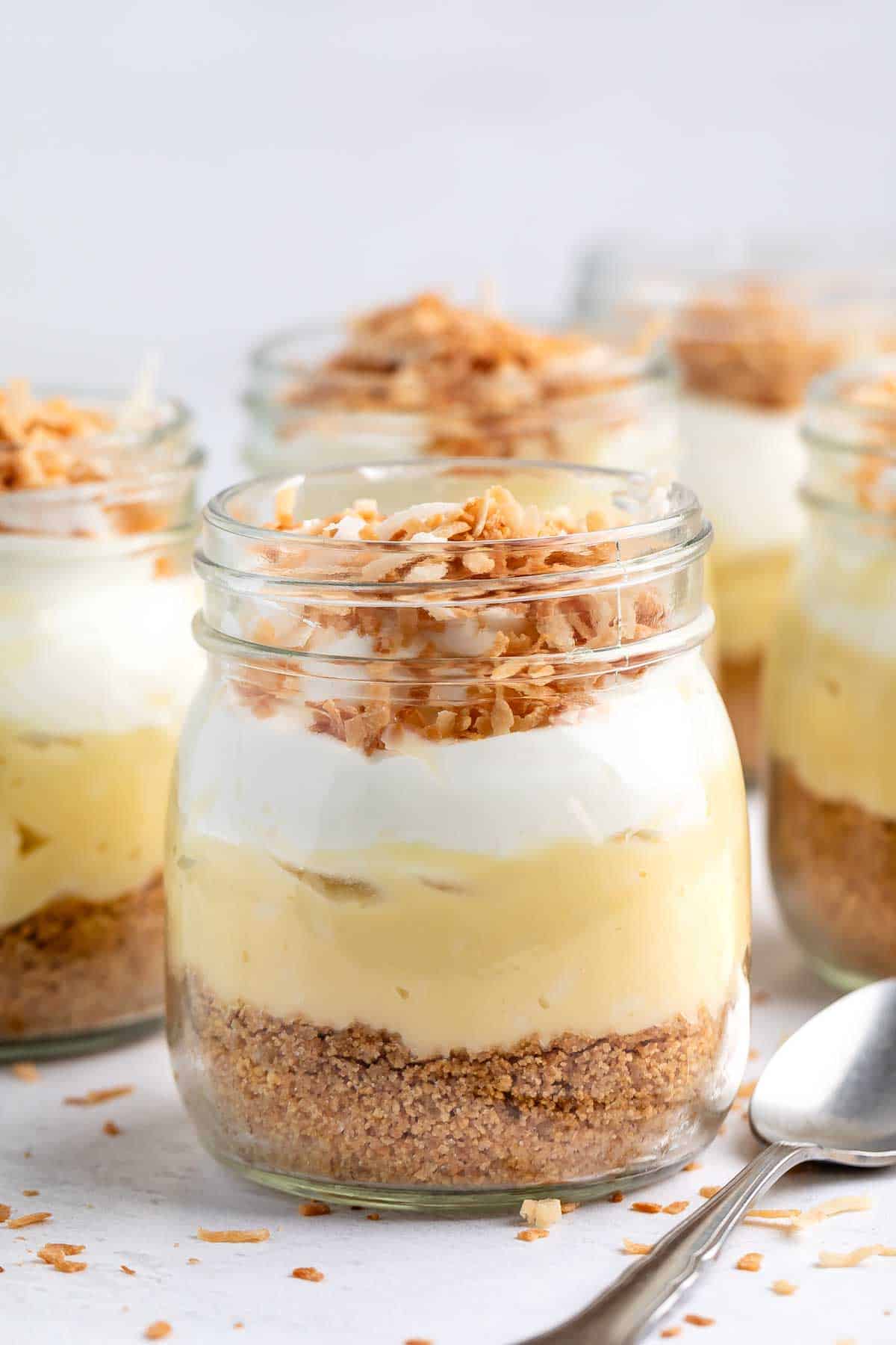 Glass jars filled with layered dessert: a base of crushed biscuits, creamy yellow filling, topped with white cream and sprinkled toasted coconut flakes. Spoons are placed nearby on a light surface.