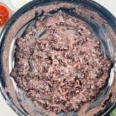 A large black bowl filled with easy refried beans sits at the center. Surrounding the bowl are small dishes containing whole black beans, sour cream, and red salsa, all on a light countertop.
