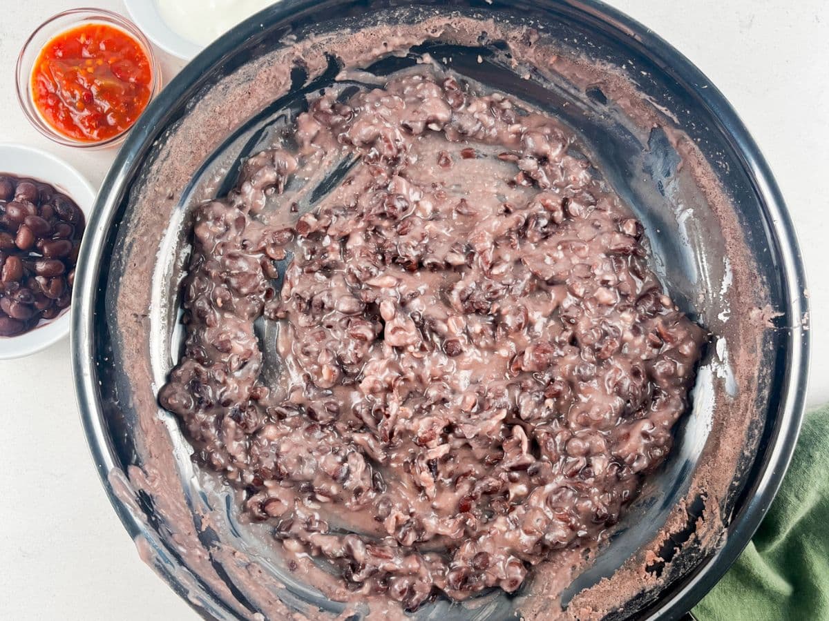 A large black bowl filled with easy refried beans sits at the center. Surrounding the bowl are small dishes containing whole black beans, sour cream, and red salsa, all on a light countertop.