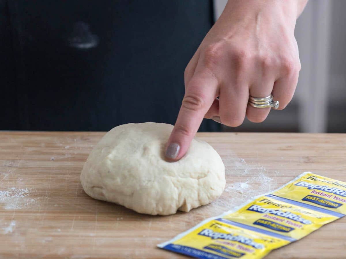 A person presses their finger into a ball of dough on a wooden surface. Three packets of instant yeast rest nearby, and a person with a dark apron is in the background. Flour is scattered on the surface.