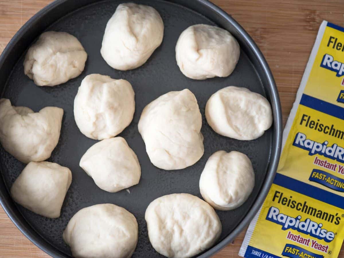 Twelve dough balls are arranged in a circular baking pan, ready for baking. Nearby, packets of Fleischmanns RapidRise Instant Yeast are visible on a wooden surface.