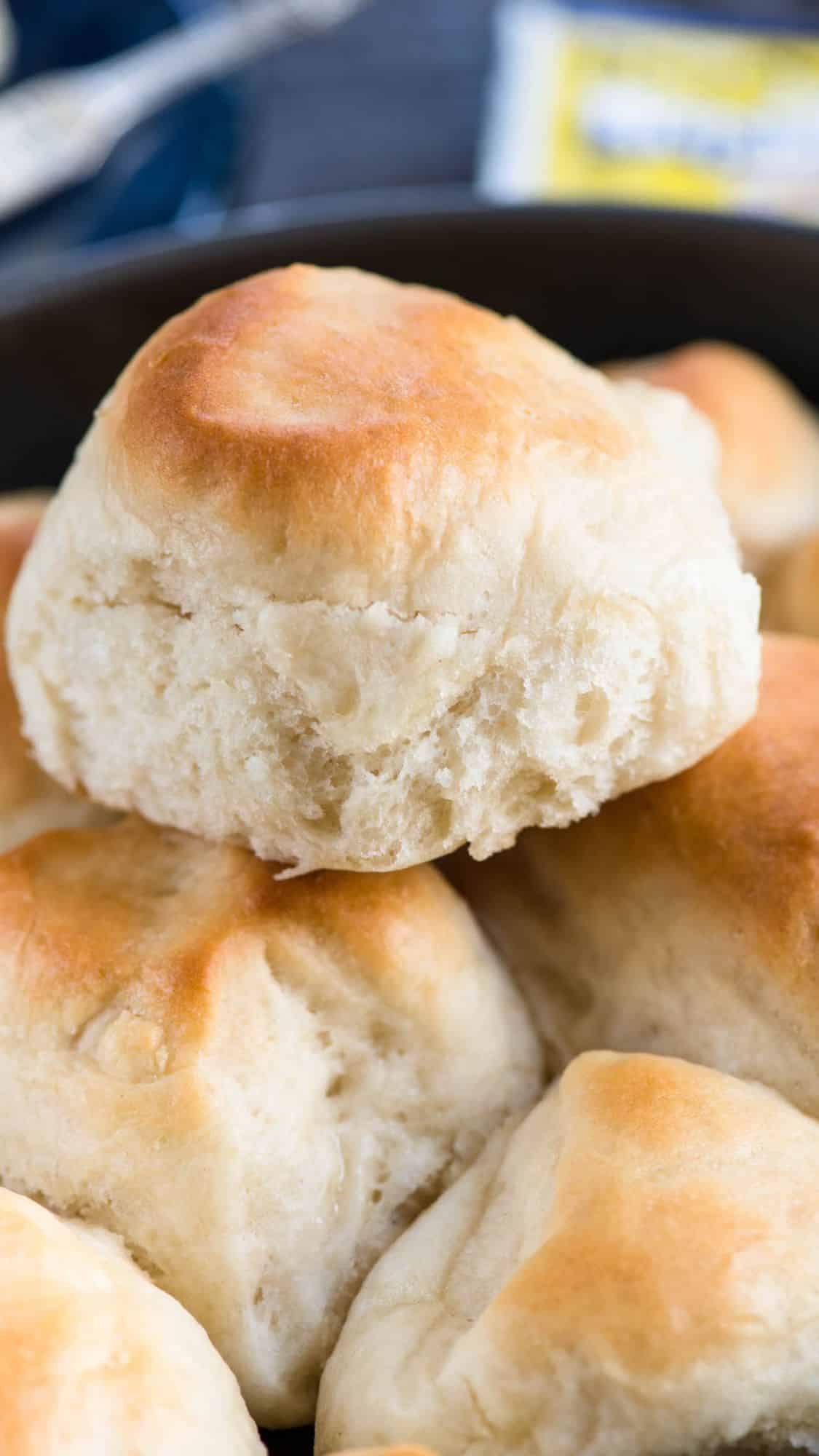 A stack of freshly baked dinner rolls with a golden-brown top. The soft, fluffy texture is visible, and they are arranged in a black dish. A blurred background suggests a table setting.