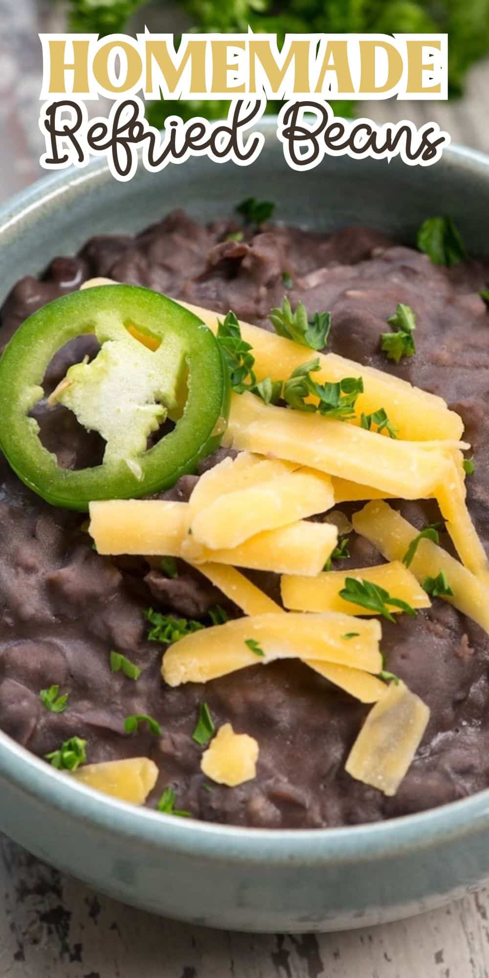 A bowl of homemade refried beans topped with sliced jalapeño, shredded cheese, and chopped herbs. Text at the top reads Homemade Refried Beans. The beans are dark, creamy, and garnished in a rustic bowl.