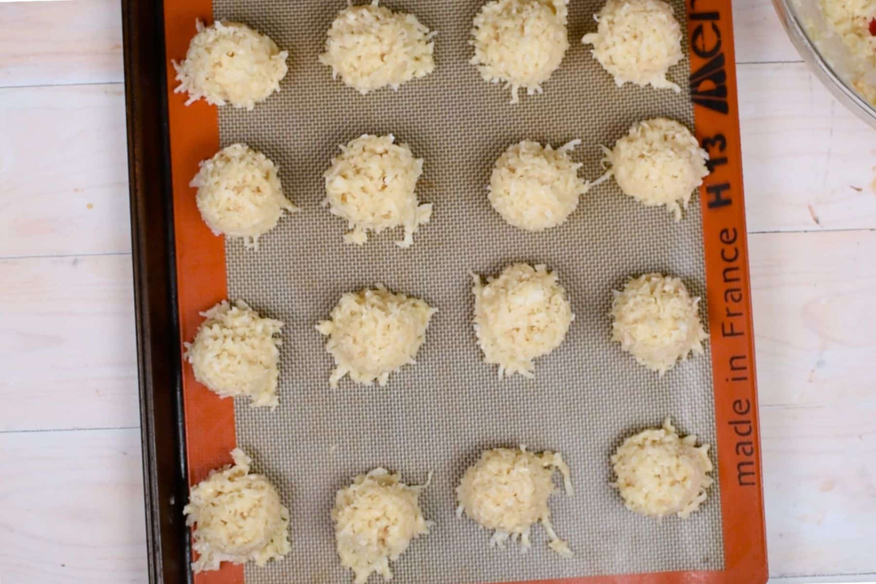 Unbaked coconut macaroons are arranged in rows on a baking sheet lined with a silicone mat. The mat is orange on the edges and labeled made in France. The surface below is light-colored wood.