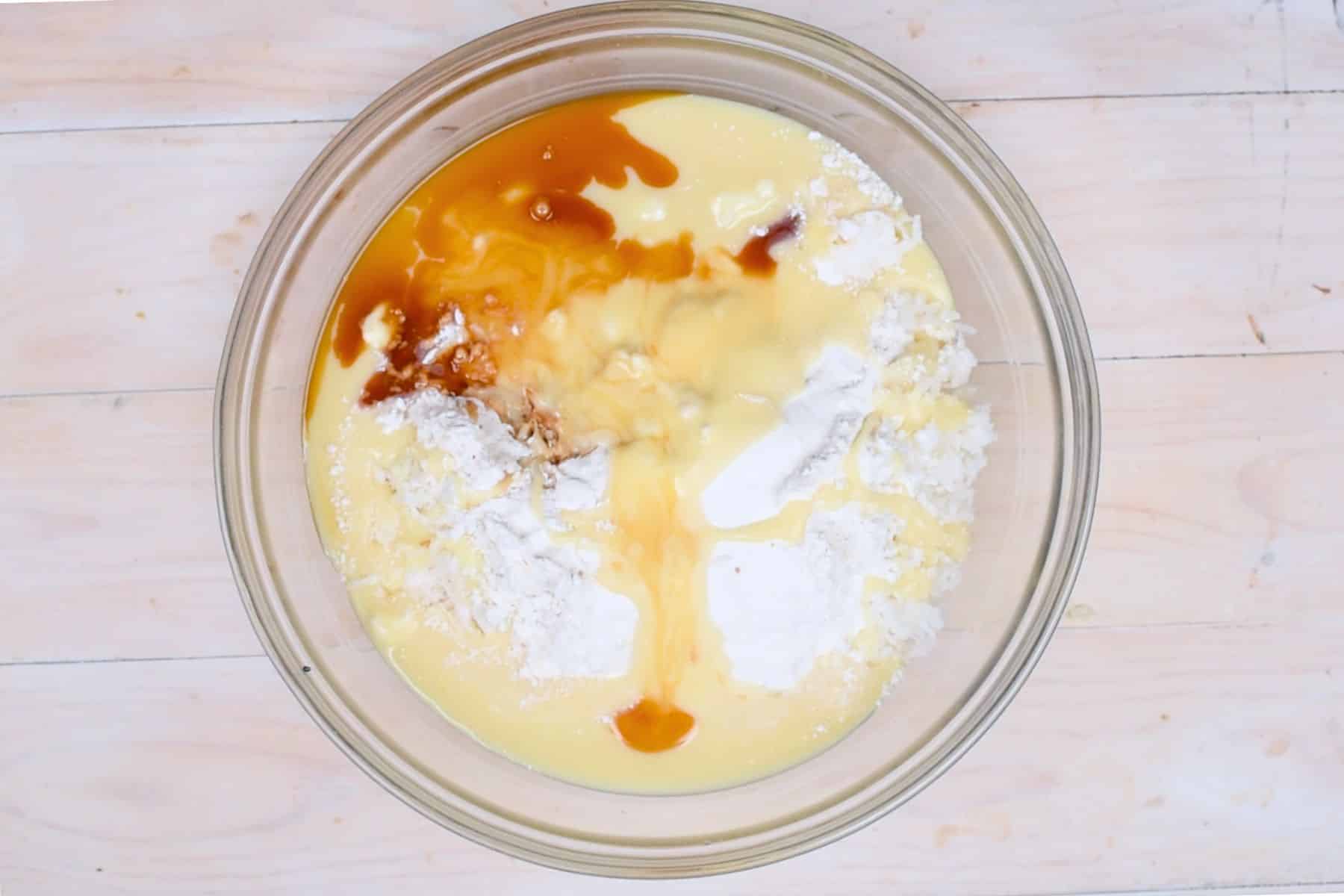 A glass mixing bowl containing flour, vanilla extract, and wet ingredients is placed on a light wooden surface. The ingredients are not yet mixed, displaying a variety of textures and colors.