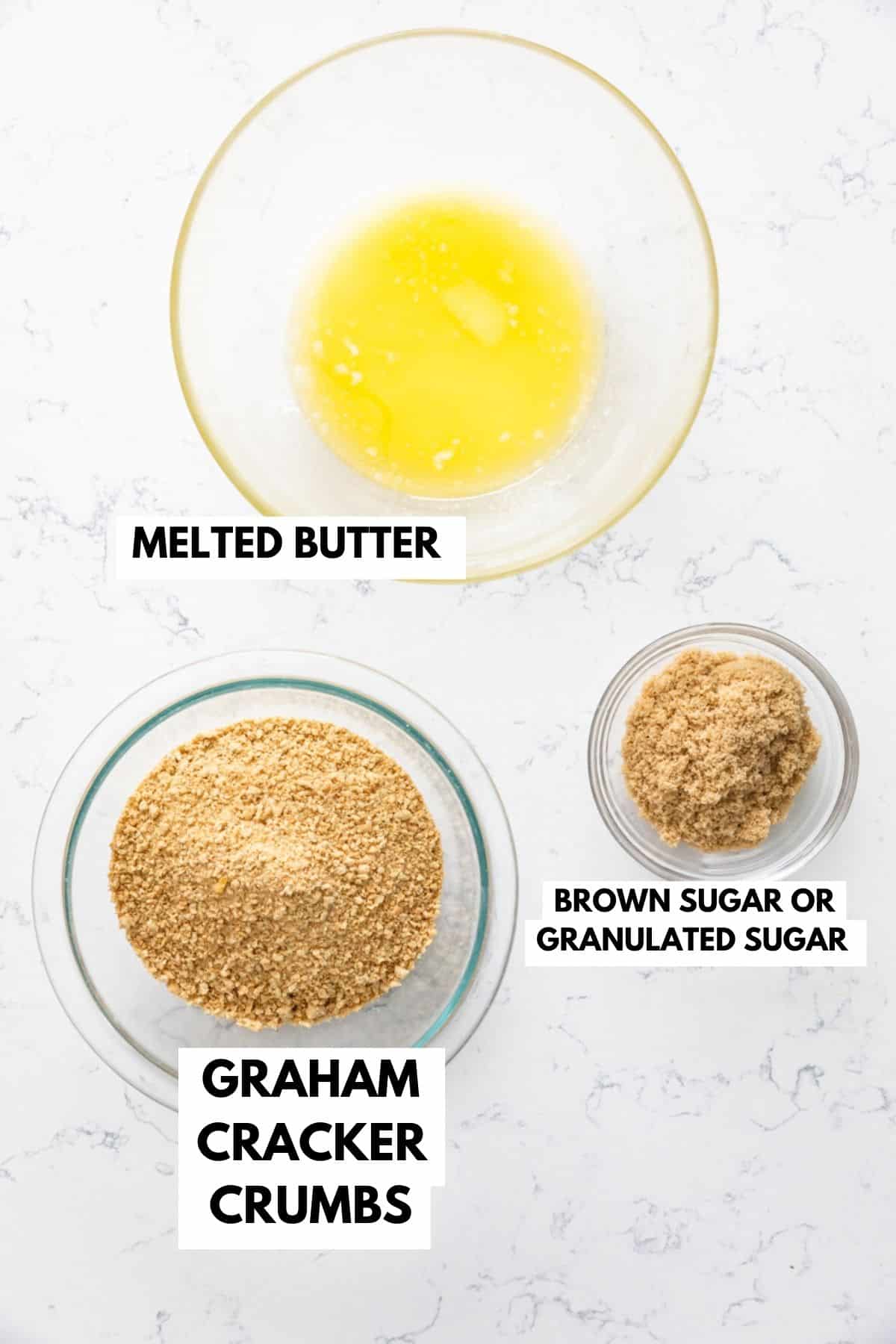 Overhead view of three bowls on a white countertop: a large bowl with melted butter, a medium bowl with graham cracker crumbs, and a small bowl with brown sugar or granulated sugar.