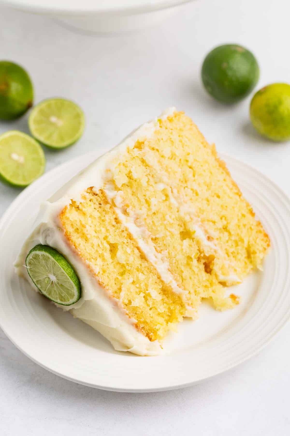 A slice of yellow citrus cake with white frosting on a white plate, garnished with a lime slice. Whole and halved limes are in the background on a light surface.