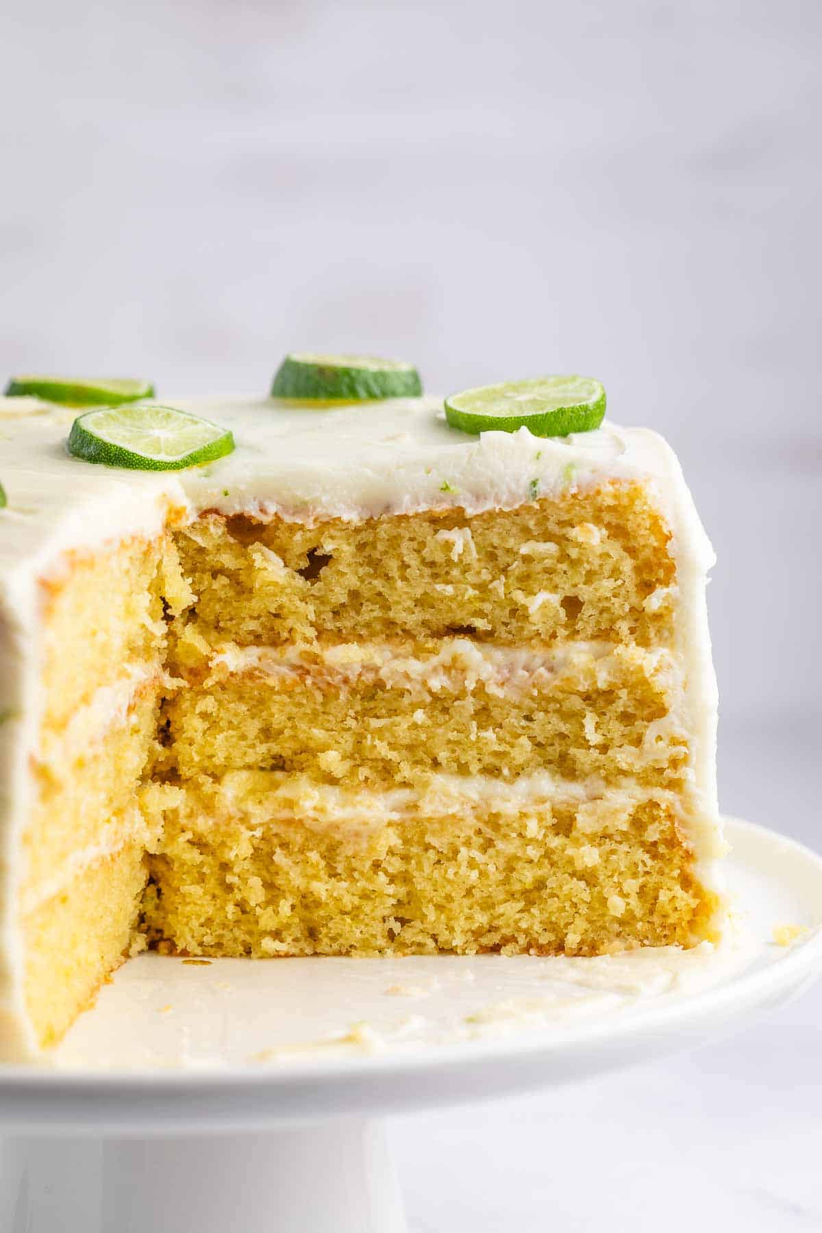 Three-layer lime cake with white frosting and lime slices on top, displayed on a white cake stand. A large slice is cut out, revealing the cakes moist texture.