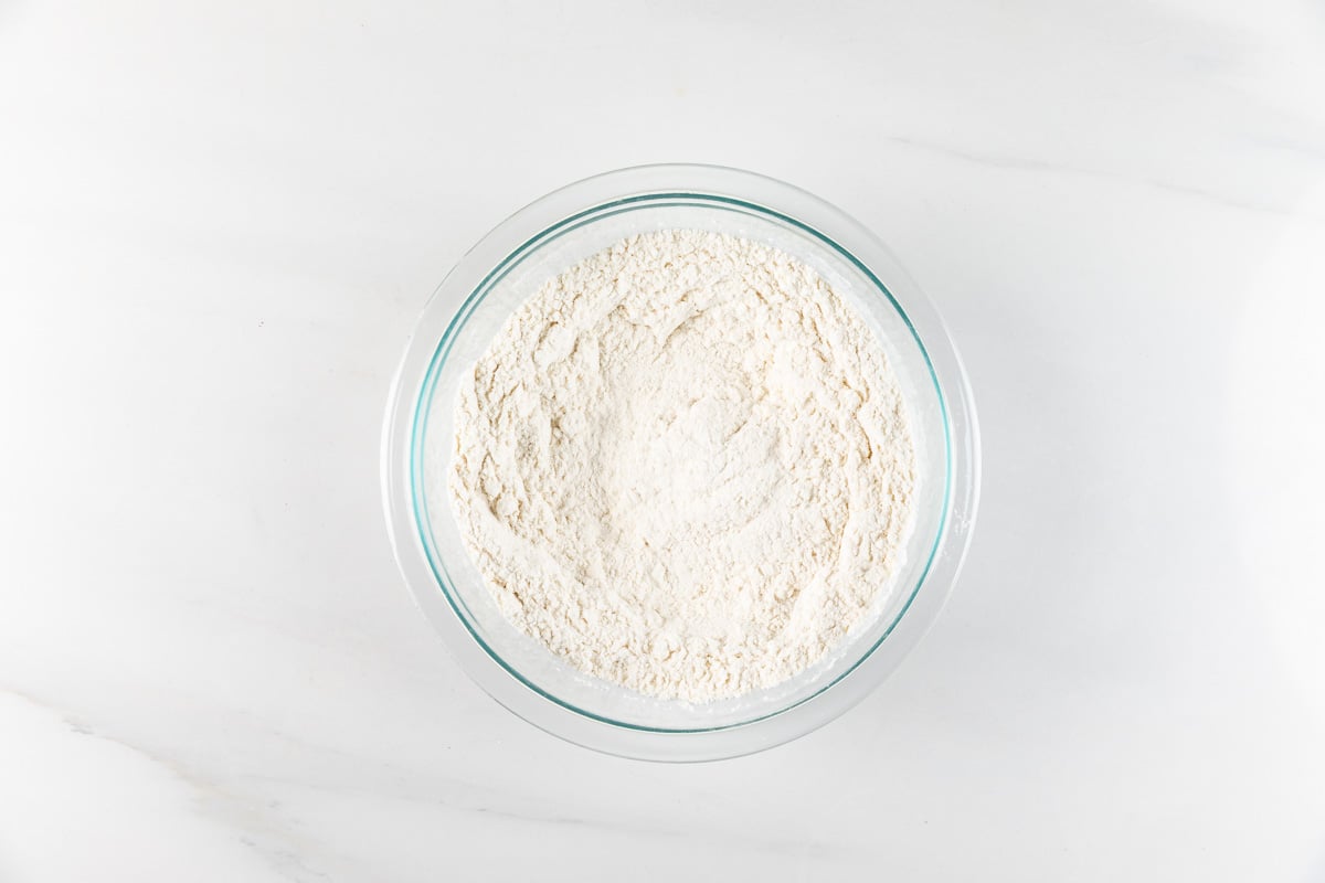 A clear glass bowl filled with white flour is placed on a smooth white marble surface. The flour is evenly distributed, creating a slightly textured appearance. The setting has a clean and minimalistic look.