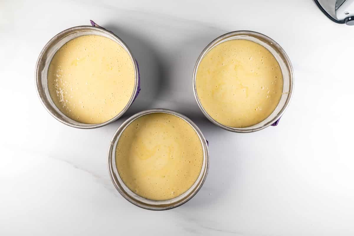 Three round cake pans filled with smooth, pale yellow batter arranged on a white surface, ready for baking.