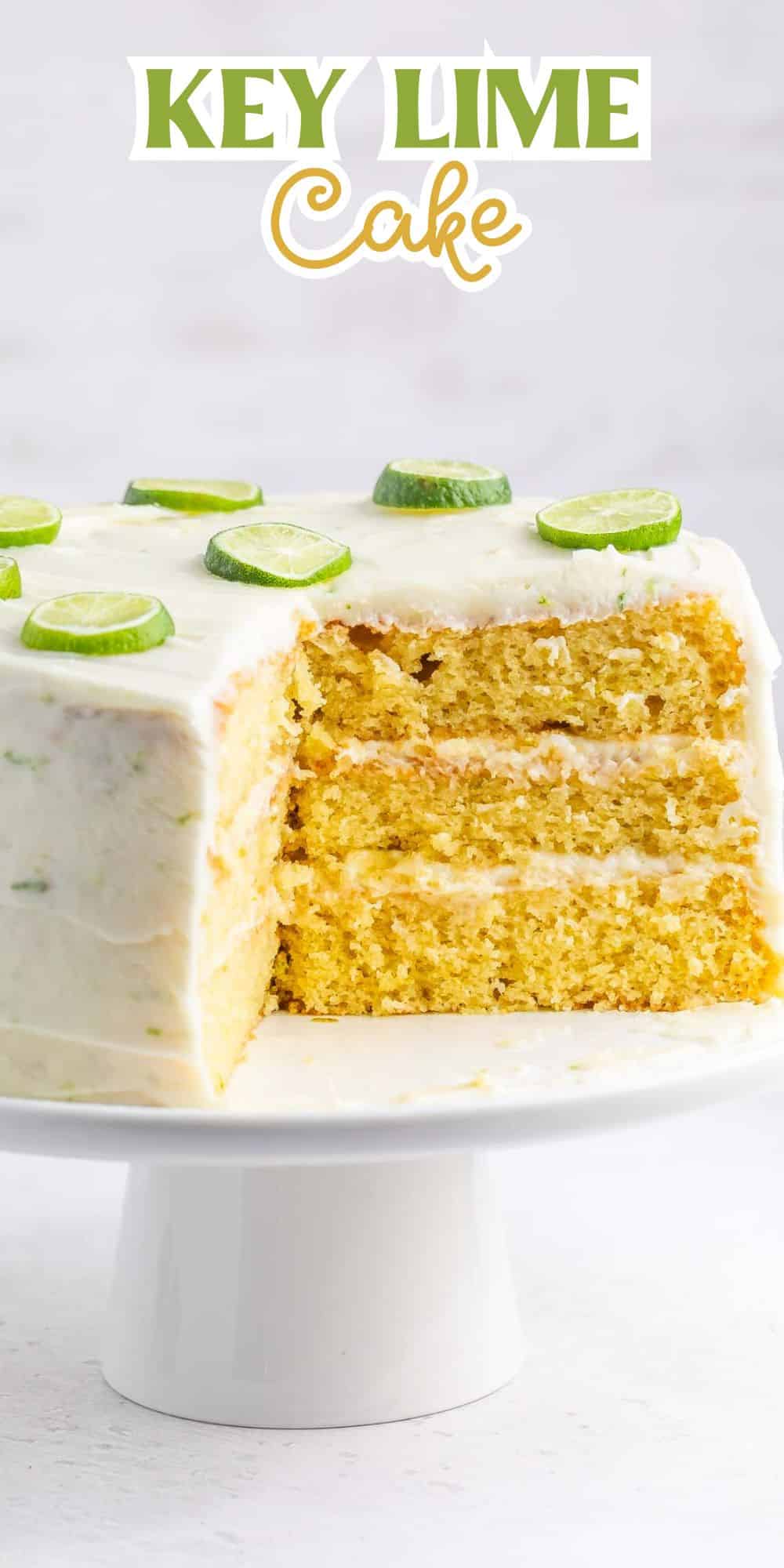 A sliced three-layer key lime cake on a white cake stand, topped with white frosting and lime slices. The background features a Key Lime Cake sign in green and yellow text.