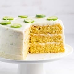 Three-layer cake with white frosting and lime slices on top, displayed on a white cake stand. A slice is cut out, revealing the moist, golden layers inside. Light background enhances the cakes vibrant colors.