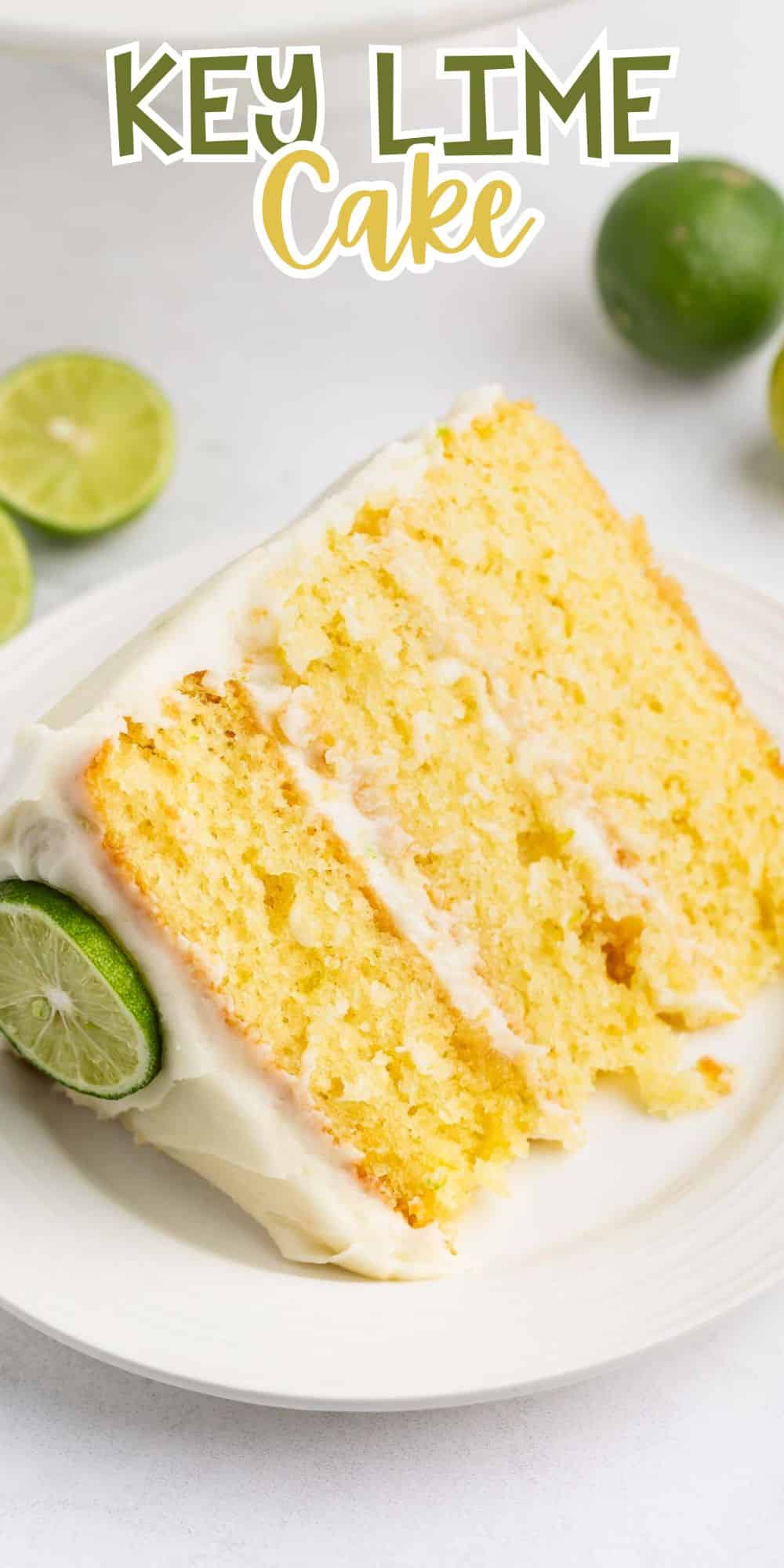A slice of key lime cake with white frosting on a plate. The cake is light yellow with visible layers, decorated with a lime slice. Whole and halved limes are in the background. The text Key Lime Cake is at the top.