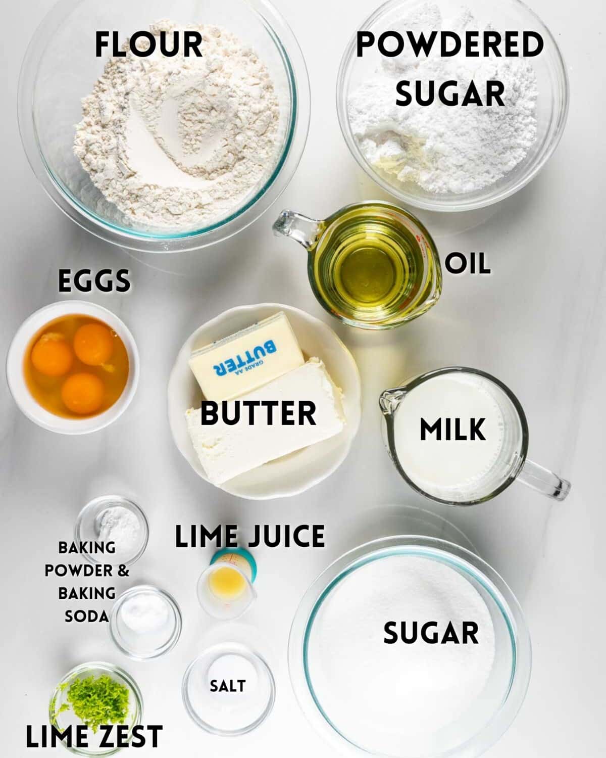 Top-down view of baking ingredients on a white surface, including flour, powdered sugar, eggs, oil, butter, milk, lime juice, sugar, lime zest, baking powder, baking soda, and salt, each in separate bowls or containers.