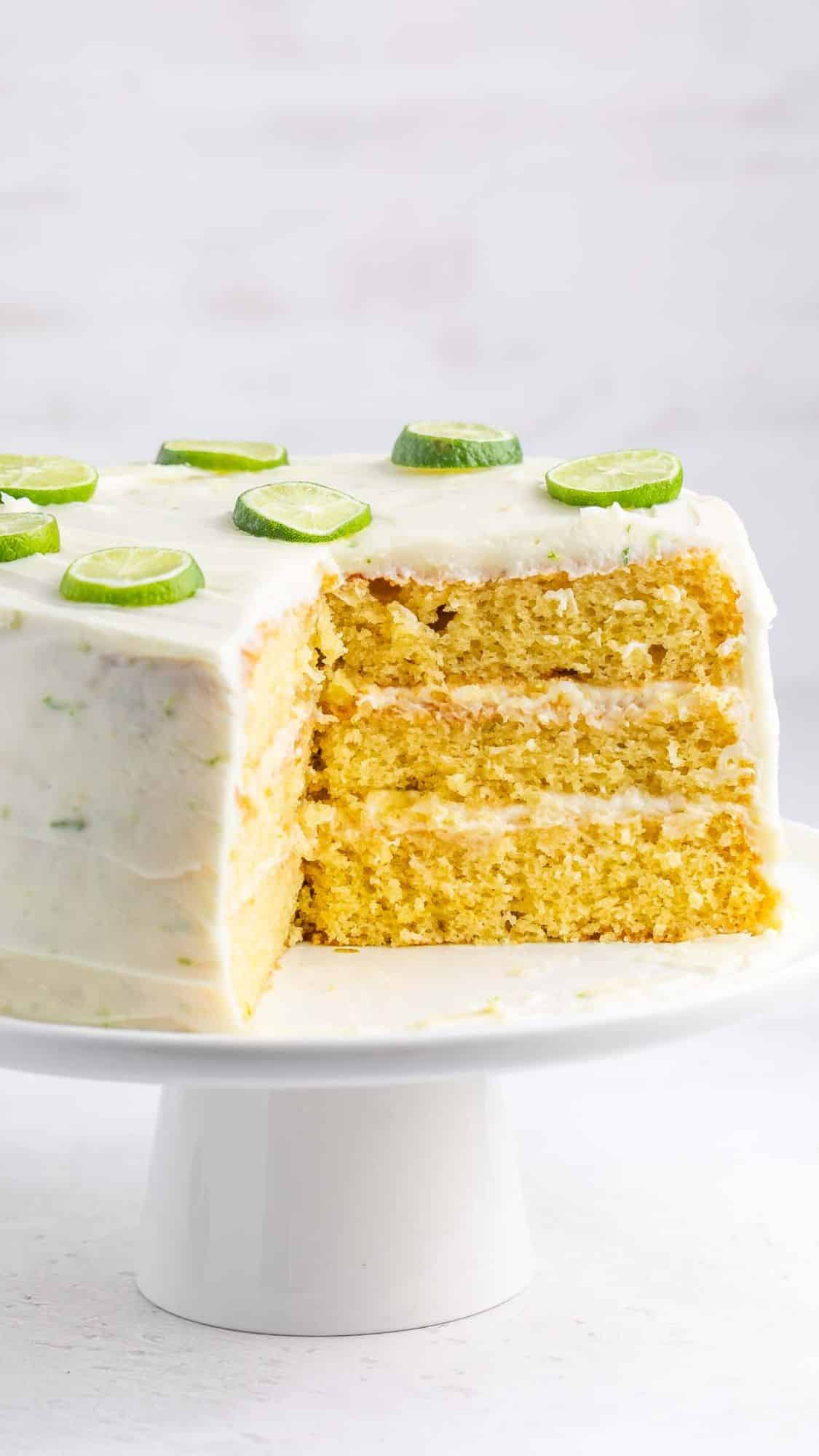 A three-layer cake with white icing, topped with lime slices, on a white cake stand. A quarter of the cake is cut out, showing the moist, yellow interior layers. The background is a soft, neutral tone.