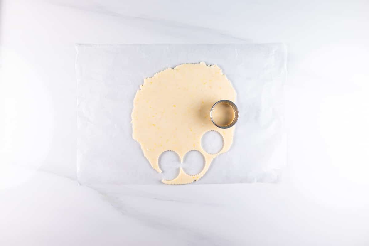 A piece of dough rolled out on parchment paper with three circular shapes cut out using a cookie cutter. The dough is placed on a light-colored, smooth surface.
