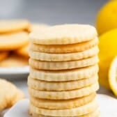 A stack of round lemon cookies on a marble surface, with a blurred background showing more cookies and a whole lemon. The cookies are light golden with visible lemon zest.