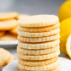 A stack of round lemon cookies on a marble surface, with a blurred background showing more cookies and a whole lemon. The cookies are light golden with visible lemon zest.
