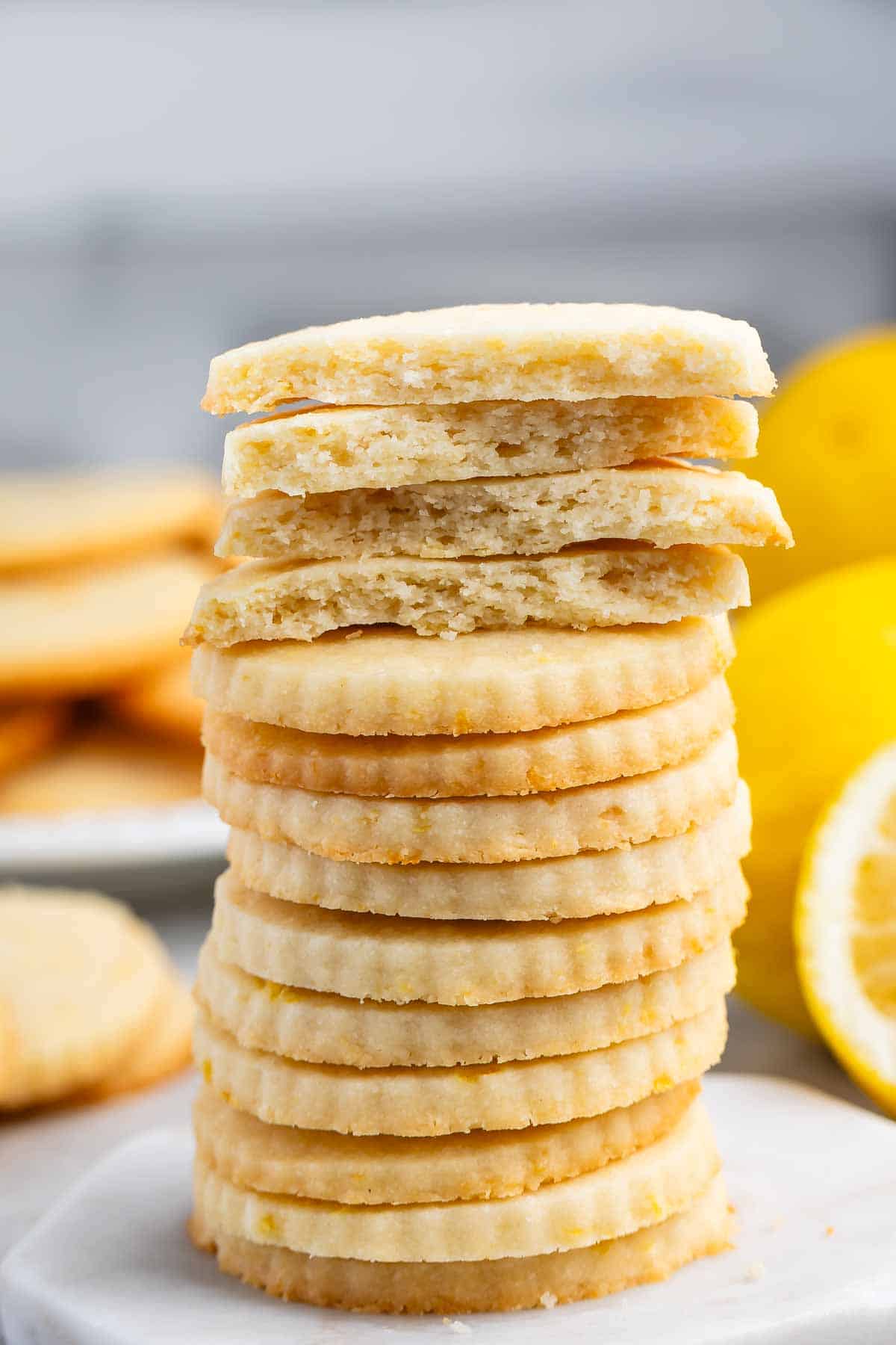 A stack of lemon cookies is neatly piled with the top few cookies broken, revealing a crumbly texture. Fresh lemons are visible in the background, enhancing the citrus theme. The setting is on a marble surface.