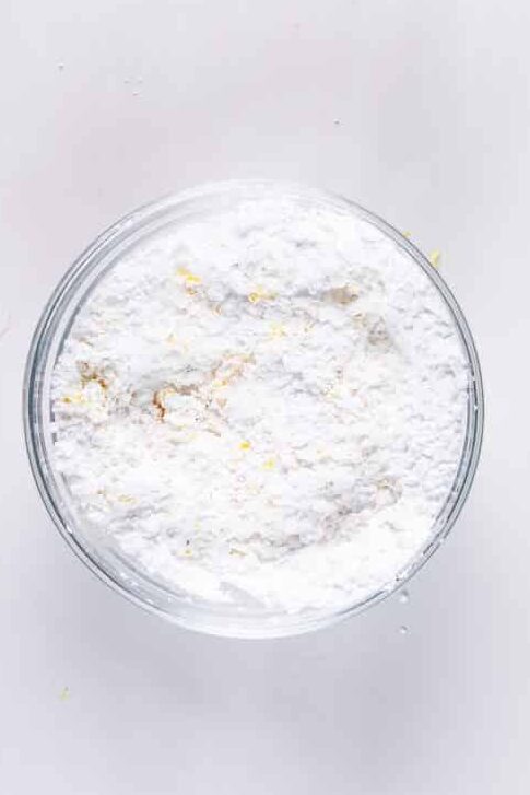 A clear glass bowl filled with white flour sits on a smooth, white surface. The flour is slightly uneven in texture, with a few small clumps visible.