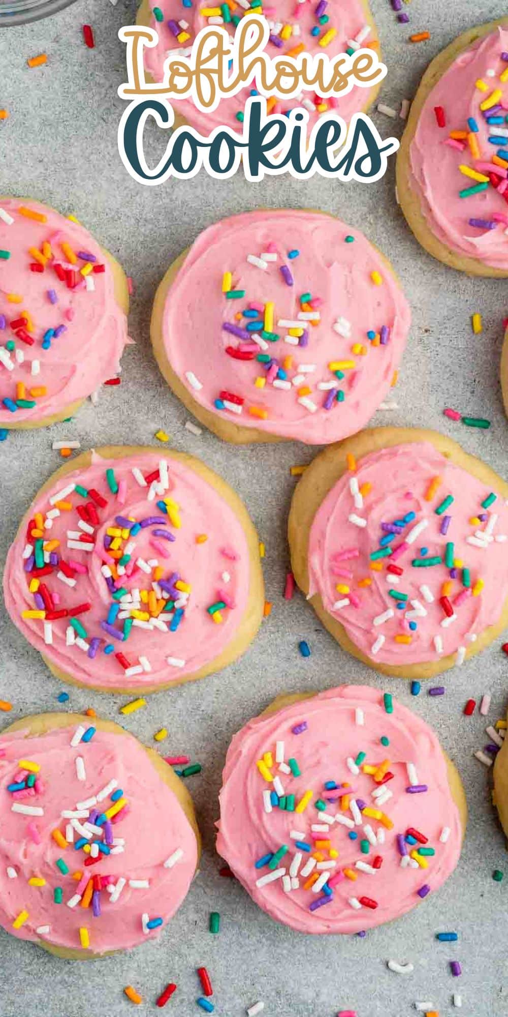 A tray of Lofthouse cookies with pink frosting and colorful sprinkles spread on a light gray surface. The cookies are soft, round, and evenly spaced, with vibrant sprinkles adding a playful touch.