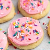 Close-up of sugar cookies topped with pink frosting and colorful sprinkles, arranged on a light-colored surface. The cookies are soft and round, with a smooth layer of icing decorated with red, yellow, blue, and purple sprinkles.