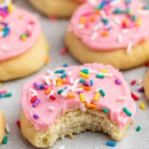 A close-up of sugar cookies with pink frosting and colorful sprinkles. One cookie in the foreground has a bite taken out, revealing a soft interior. The cookies are arranged on a light gray surface.