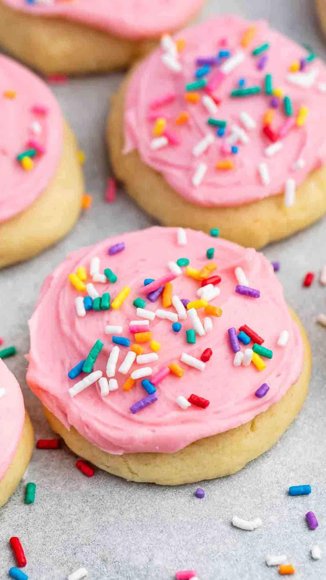 Close-up of sugar cookies topped with pink frosting and multicolored sprinkles on a light gray surface. The cookies have a soft, round appearance with a smooth layer of icing. Some sprinkles are scattered around the cookies.