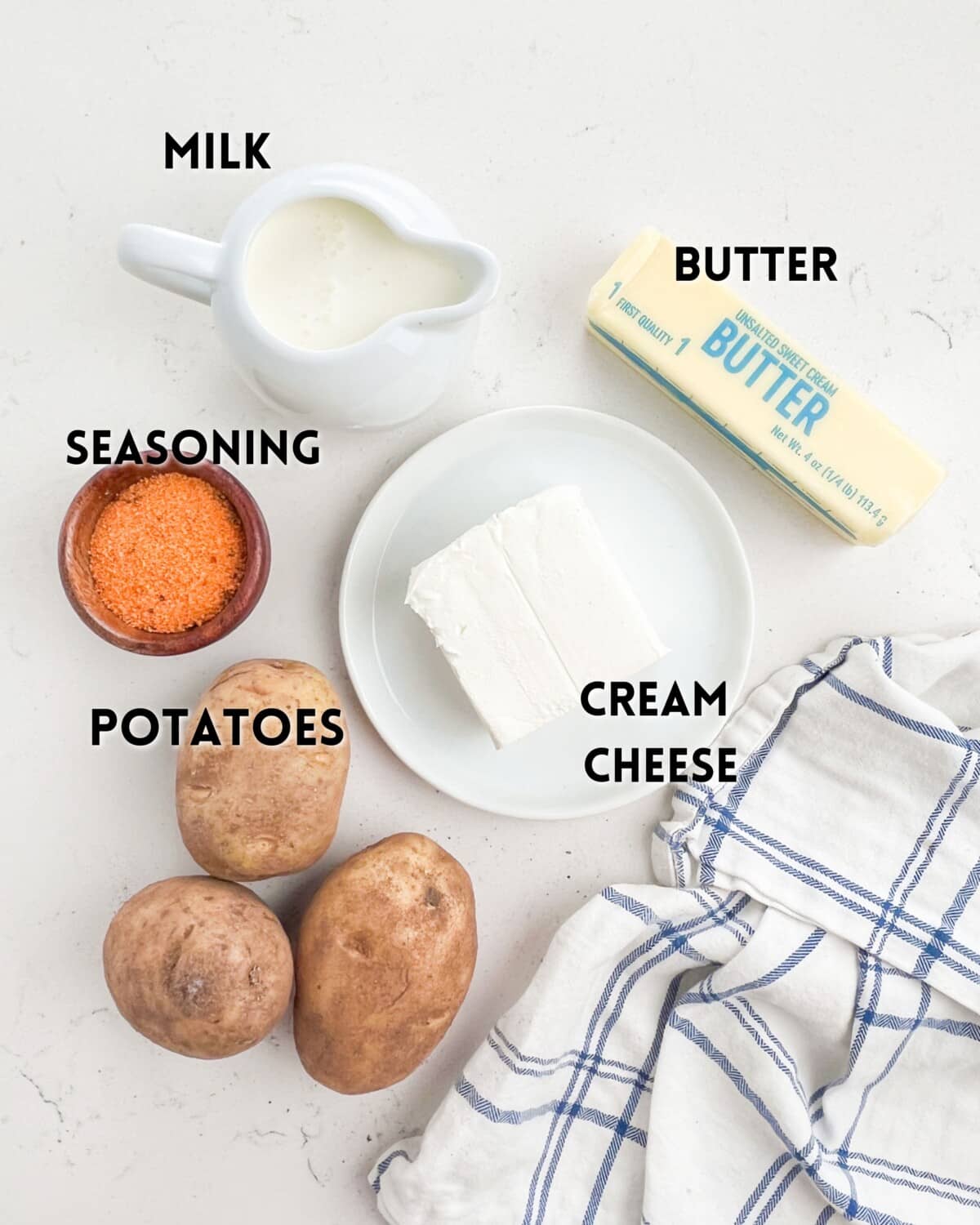 Ingredients for mashed potatoes: a jug of milk, a stick of butter, a chunk of cream cheese on a plate, a bowl of seasoning, three potatoes, and a striped towel, arranged on a light surface.