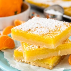 Three lemon bars stacked on a light blue plate, dusted with powdered sugar. In the background, there are sliced oranges, striped straws, and a small bowl of sugar on a white surface.