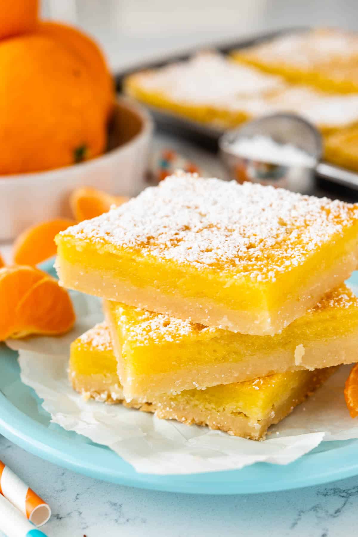 Three lemon bars stacked on a light blue plate, dusted with powdered sugar. In the background, there are sliced oranges, striped straws, and a small bowl of sugar on a white surface.