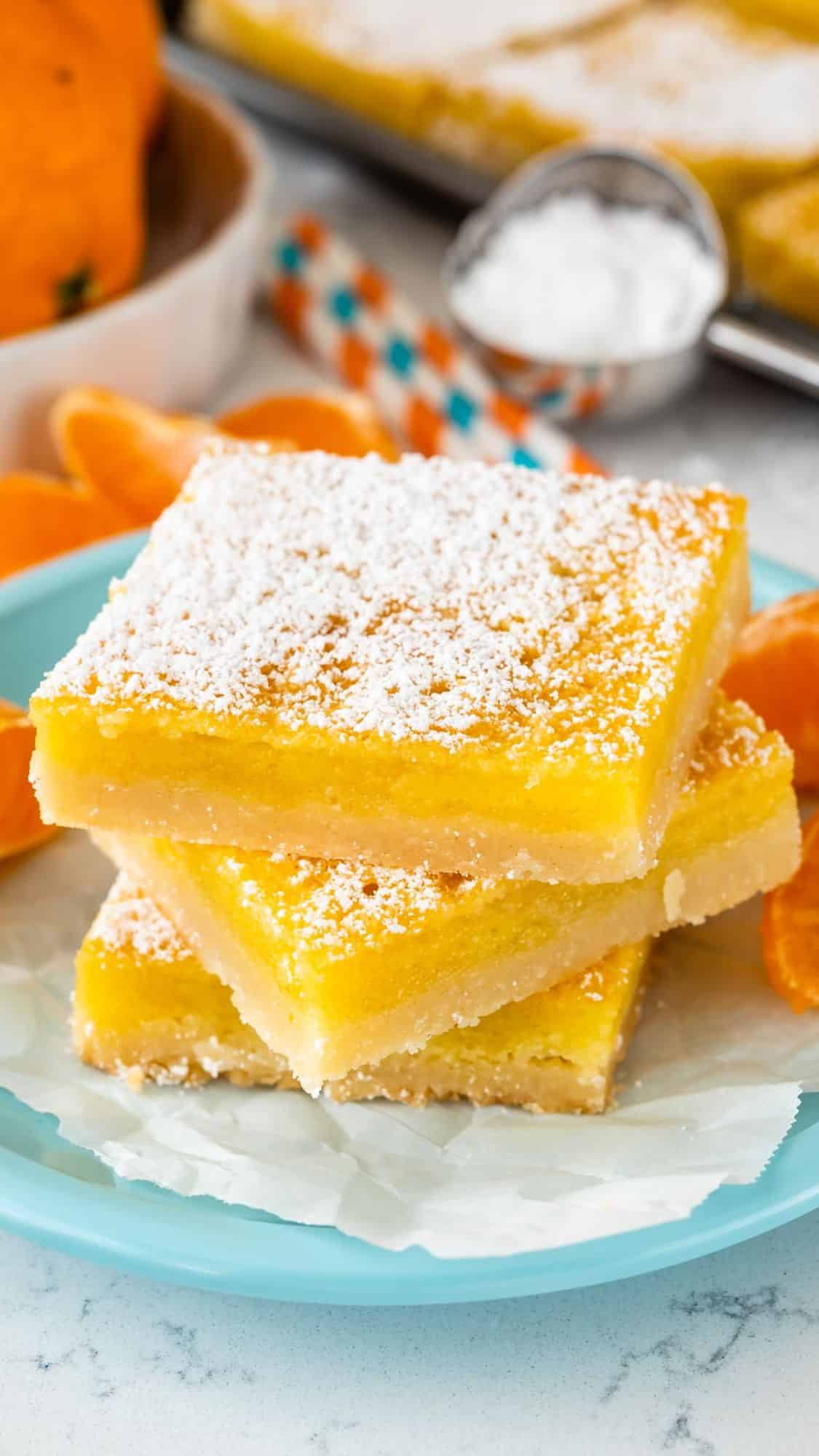 Three lemon bars stacked on a light blue plate, dusted with powdered sugar. In the background, there are sliced oranges, striped straws, and a small bowl of sugar on a white surface.
