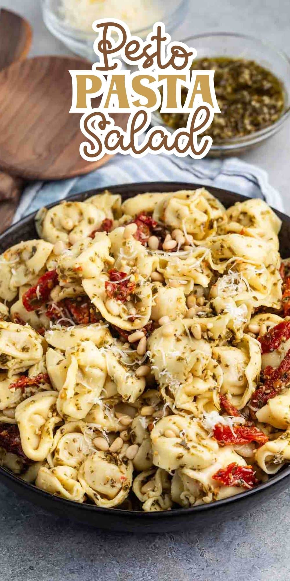 A dish of pesto pasta salad with tortellini, sun-dried tomatoes, and pine nuts in a black bowl. A bowl of pesto and grated cheese are in the background. Text overlay reads Pesto Pasta Salad.