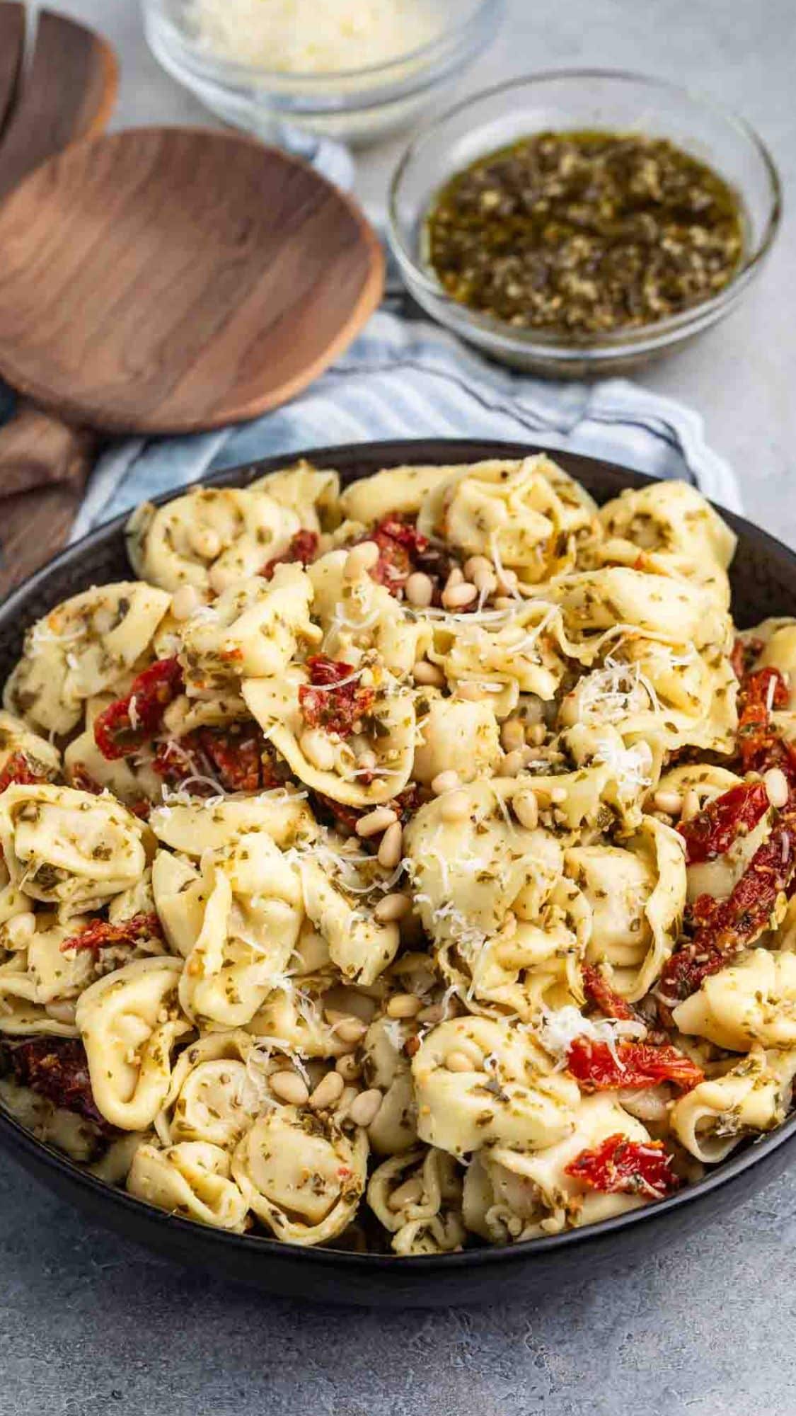 A bowl of cheese tortellini pasta covered in basil pesto sauce, garnished with sun-dried tomatoes and pine nuts. Behind the bowl, there are wooden spoons and small bowls with grated cheese and extra pesto sauce.